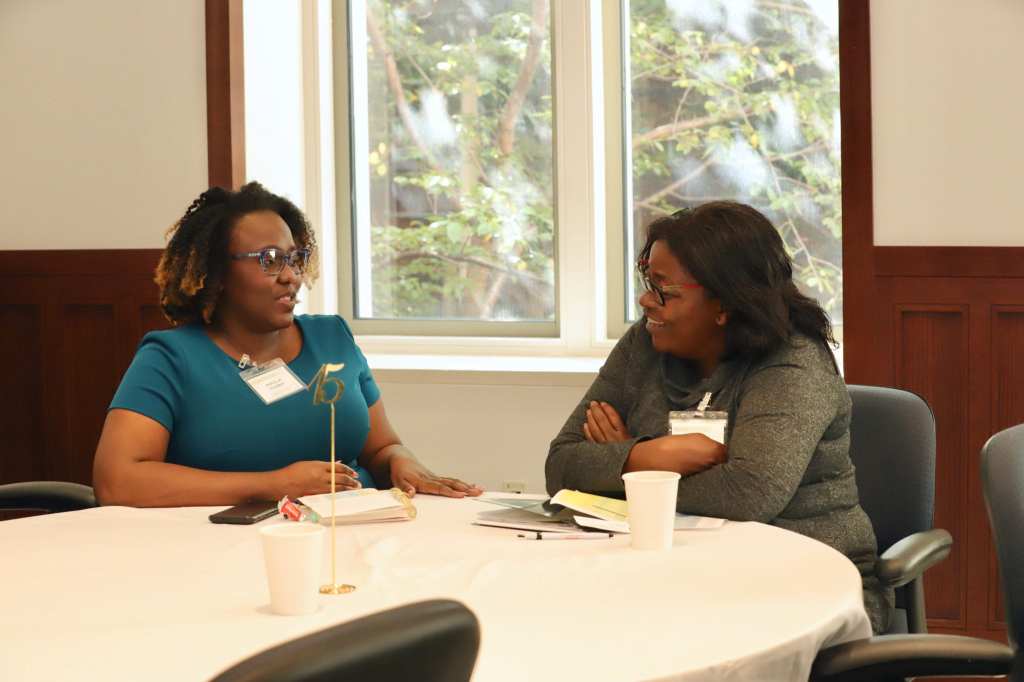 Makella sits at a conference banquet table with another black researcher. They're talking and smiling. In front of them on the table are cups of coffee and open notebooks. Both women are wearing conference badges.