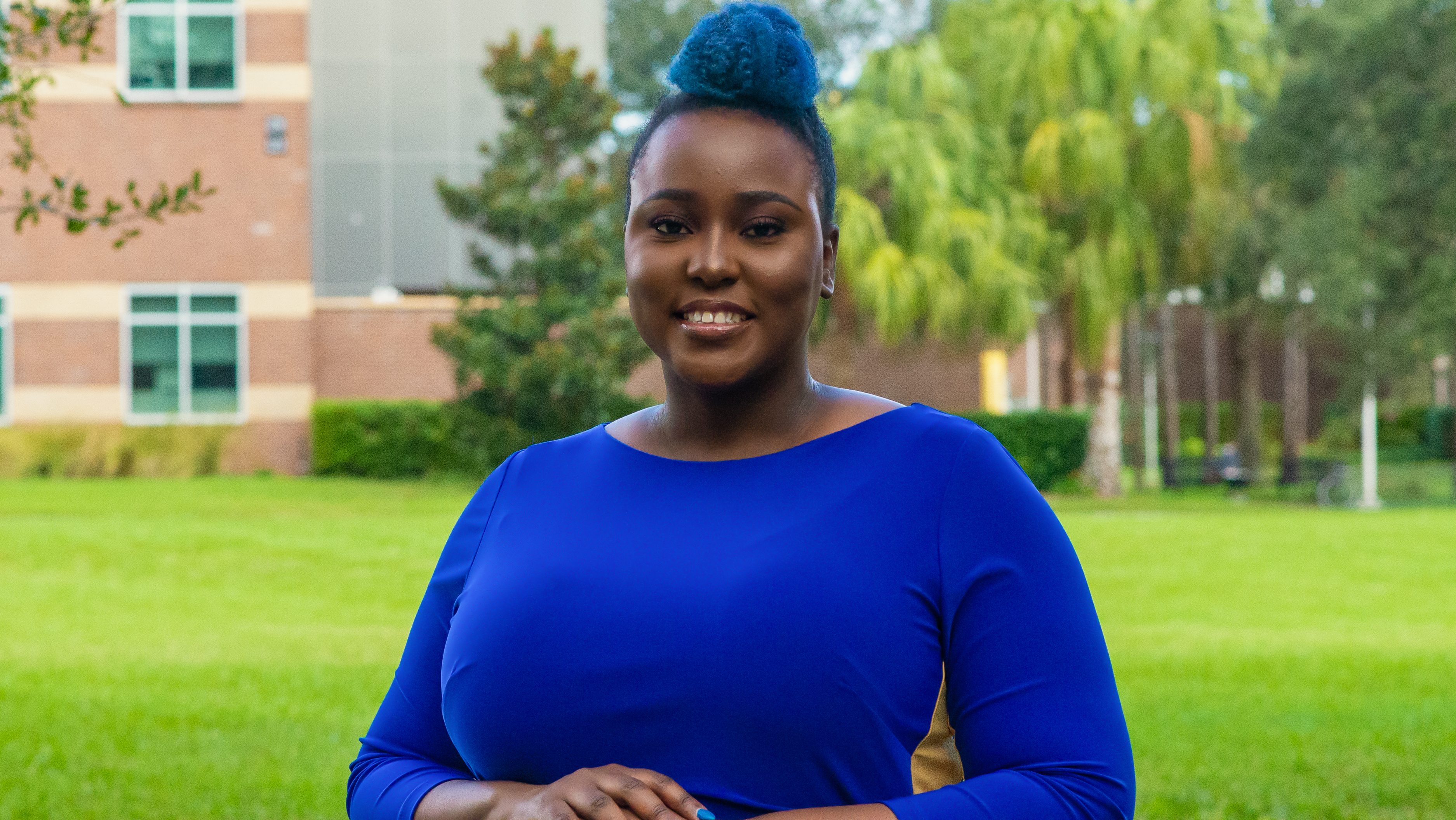 Dr. Makella Coudray smiles at the camera. She's a black woman wearing a bright blue long sleeve top that matches her blue hair which is in a bun at the top of her head. She looks cool and approachable.