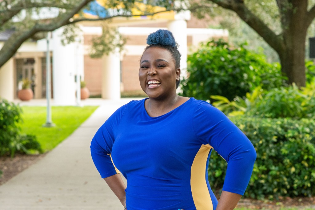 Dr. Makella Coudray smiles while laughing outside on a path at the University of Central Florida Medicine campus. Her hands are on her hips. She's wearing a blue dress with yellow stripe. The blue matches her blue hair in a bun at the top of her head.
